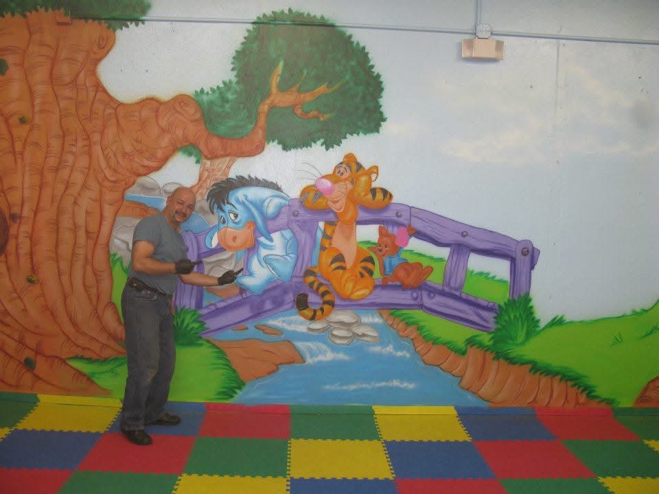 Efrain standing beside a hand-airbrushed Winnie the Pooh mural with Eeyore and Tigger on a bridge